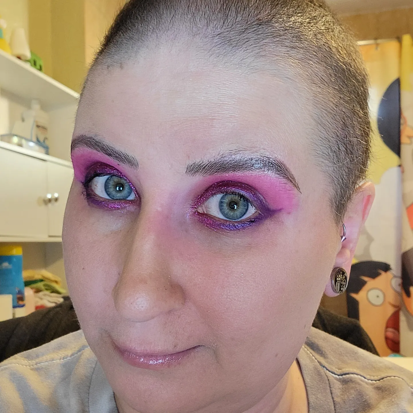A close up of a woman with very very short hair and bright pink makeup, smiling for the picture.