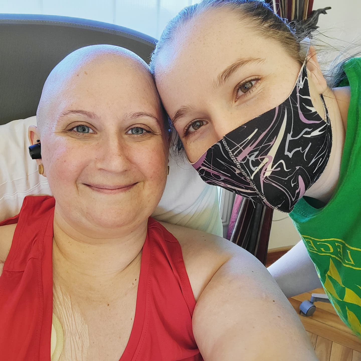 A picture of two women, one bald and in a red tank top, the other in a green shirt and a mask smiling for the picture during an infusion room.