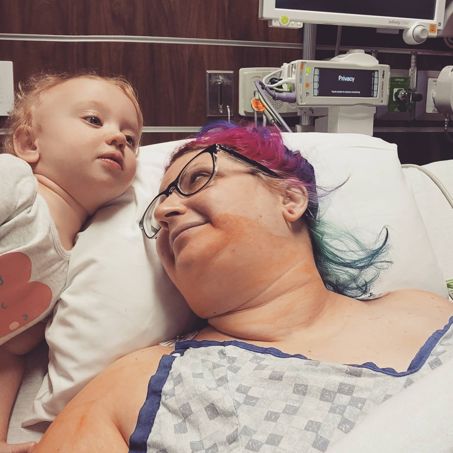 A girl just waking up from a double mastectomy surgery, lying in a hospital bed, smiling at her baby girl.