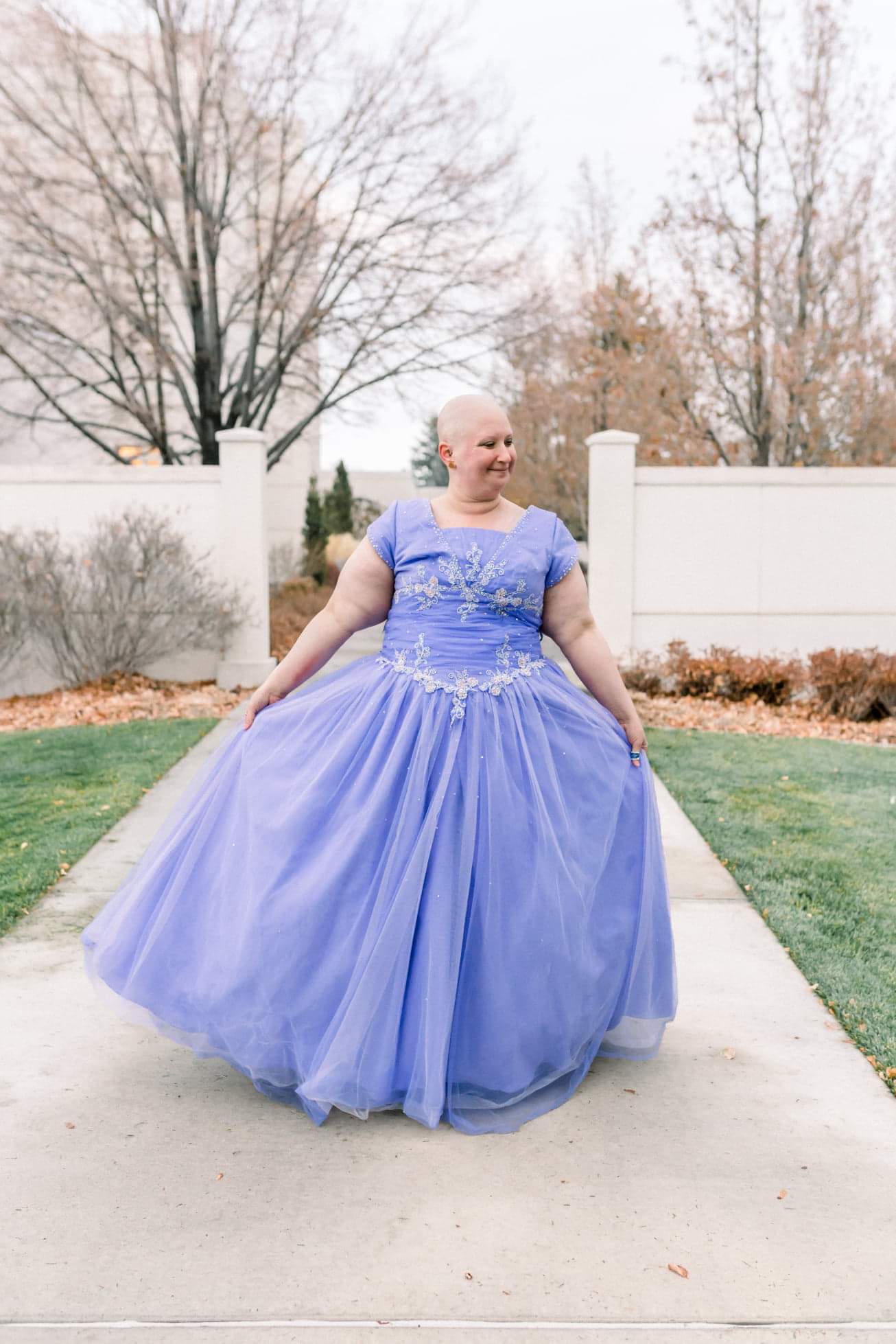 Cancer survivor walking down the sidewalk towards the camera in a purple flowy gown with a bald head.